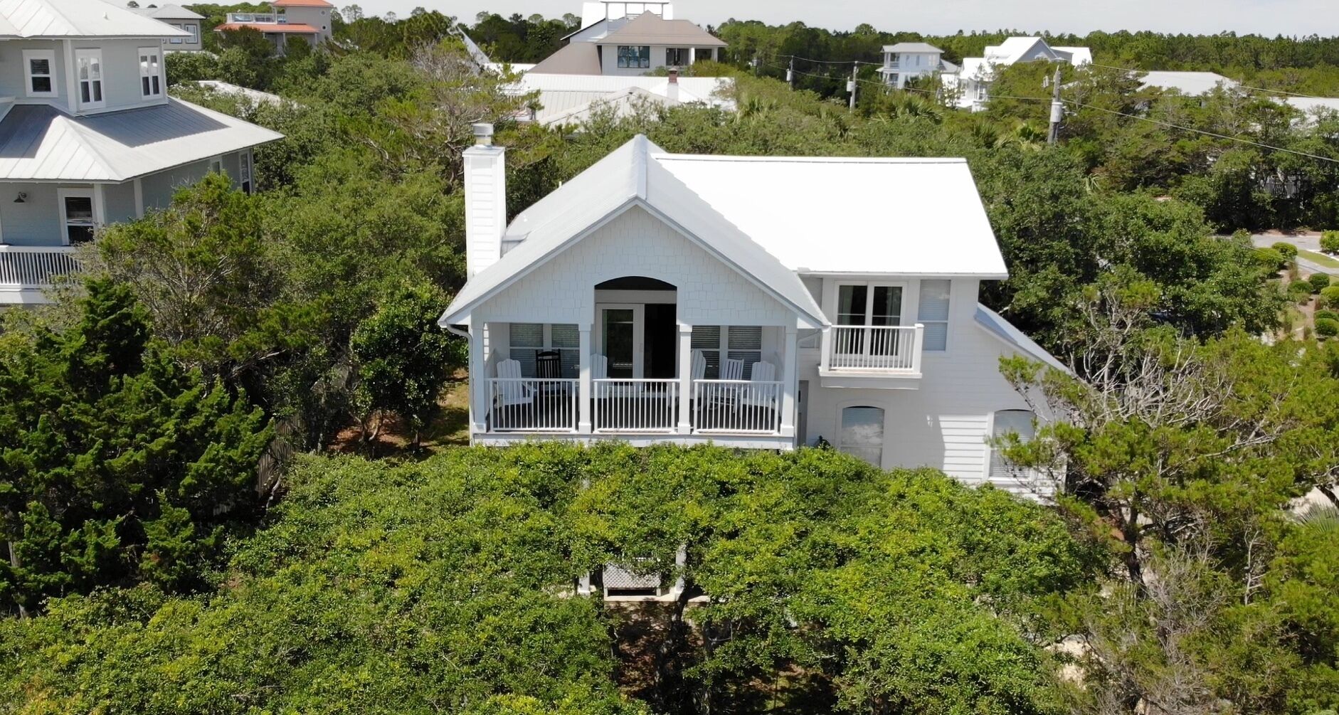 A white two-story house with a balcony, surrounded by dense greenery.
