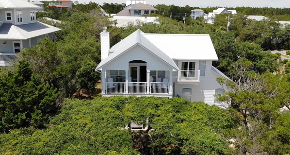 A white two-story house with a balcony, surrounded by dense greenery.