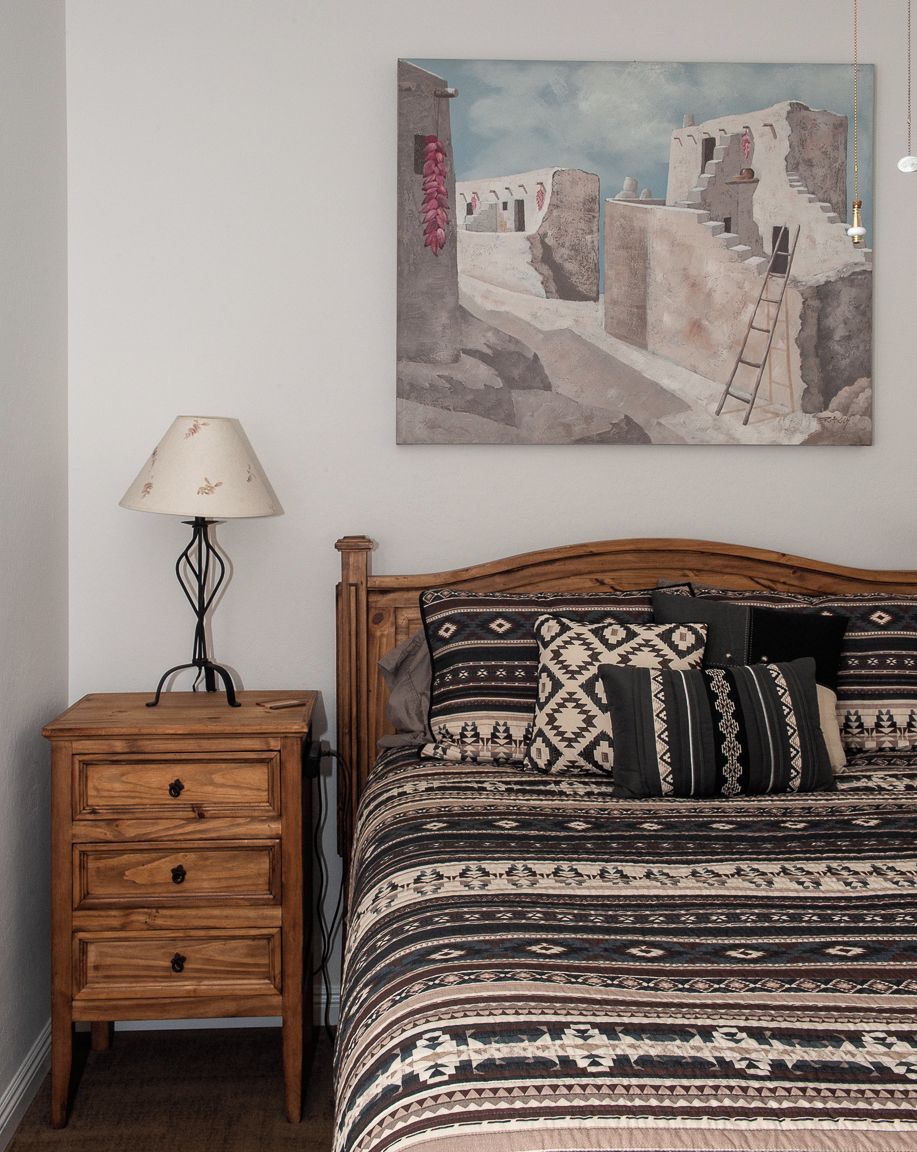 Un dormitorio con cama de madera, ropa de cama estampada, una mesita de noche con lámpara y un cuadro en la pared.