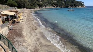 A beach with clear waters, a row of sunbeds, and a building with outdoor seating. - Furnished 3 STARS Golf St Tropez RAYOL CANADEL Facing the SEA with POOL (Rayol-Canadel-sur-Mer)