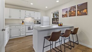 Une cuisine moderne avec des meubles blancs, un îlot central doté de tabourets et trois tableaux encadrés accrochés au mur.