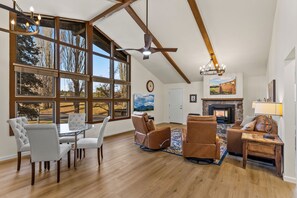 Uma sala de estar espaçosa com lareira, poltronas de couro, mesa de jantar e grandes janelas com vista para árvores e um céu limpo.