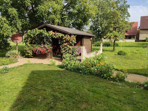 A small wooden structure with a sloped roof, surrounded by a garden with various flowering plants and a paved path leading to it. - Living like home😃 (Weigelshofen)