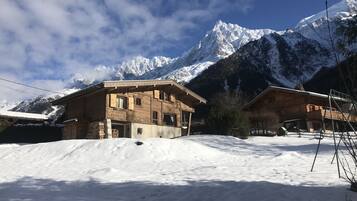 A wooden chalet with a snow-covered roof, set against a backdrop of snow-capped mountains and a clear sky.