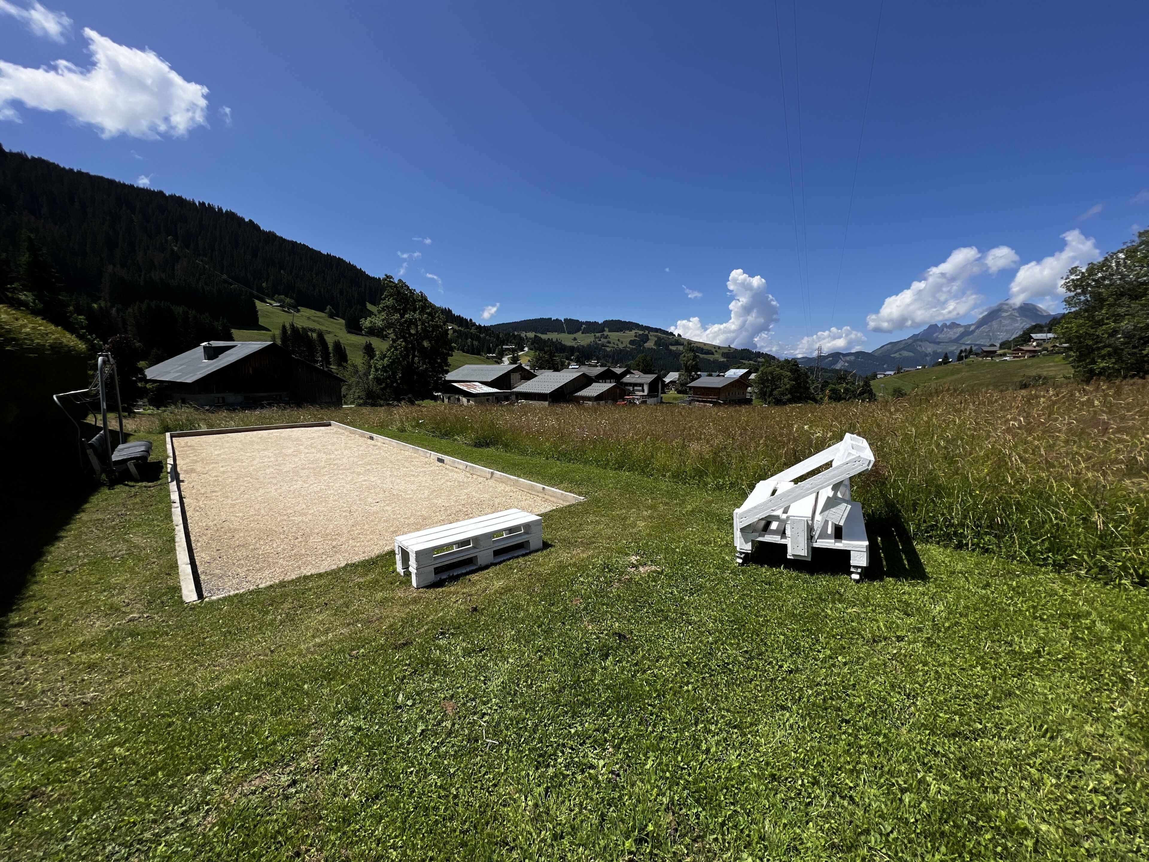A grassy field with a wooden bench and a white structure, likely a seating area, with a mountain backdrop and houses in the distance.