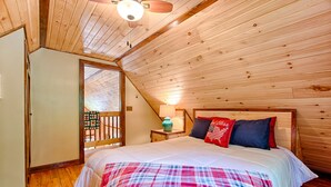 Ein Schlafzimmer mit Holzboden, einem Bett mit kariertem Überwurf, zwei Kissen, einem Nachttisch mit Lampe und Blick ins Freie durch ein Fenster.