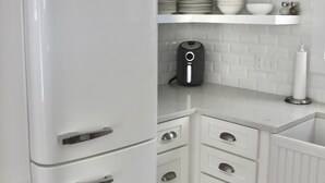 Une cuisine avec un réfrigérateur Smeg blanc, des meubles de rangement blancs, un plan de travail avec une machine à café, et des étagères où sont rangés de la verrerie et de la vaisselle.