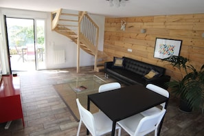 A modern living room with a glass coffee table, a black sofa, a wooden staircase, and a view of the outdoors.