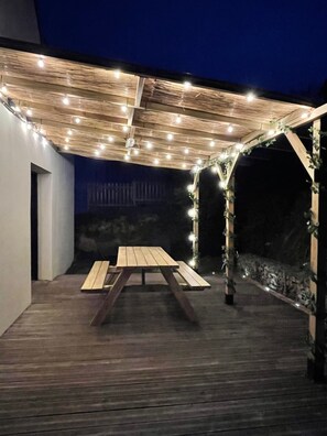 Une terrasse extérieure avec une table et des bancs en bois, des guirlandes lumineuses suspendues au-dessus et un mur en pierre.