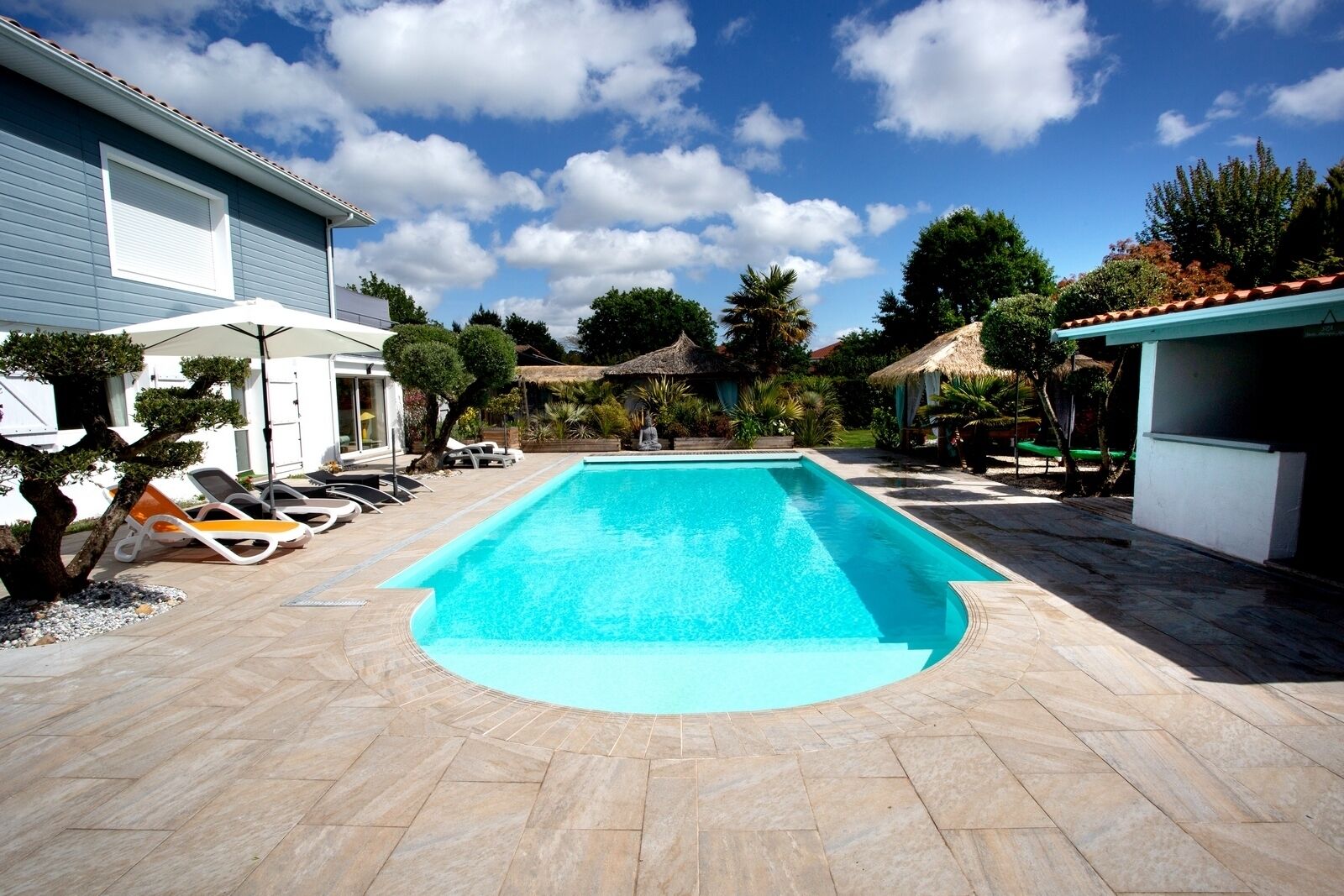 Une piscine rectangulaire, bordée d’une terrasse carrelée, entourée de chaises longues et d’un petit arbre.