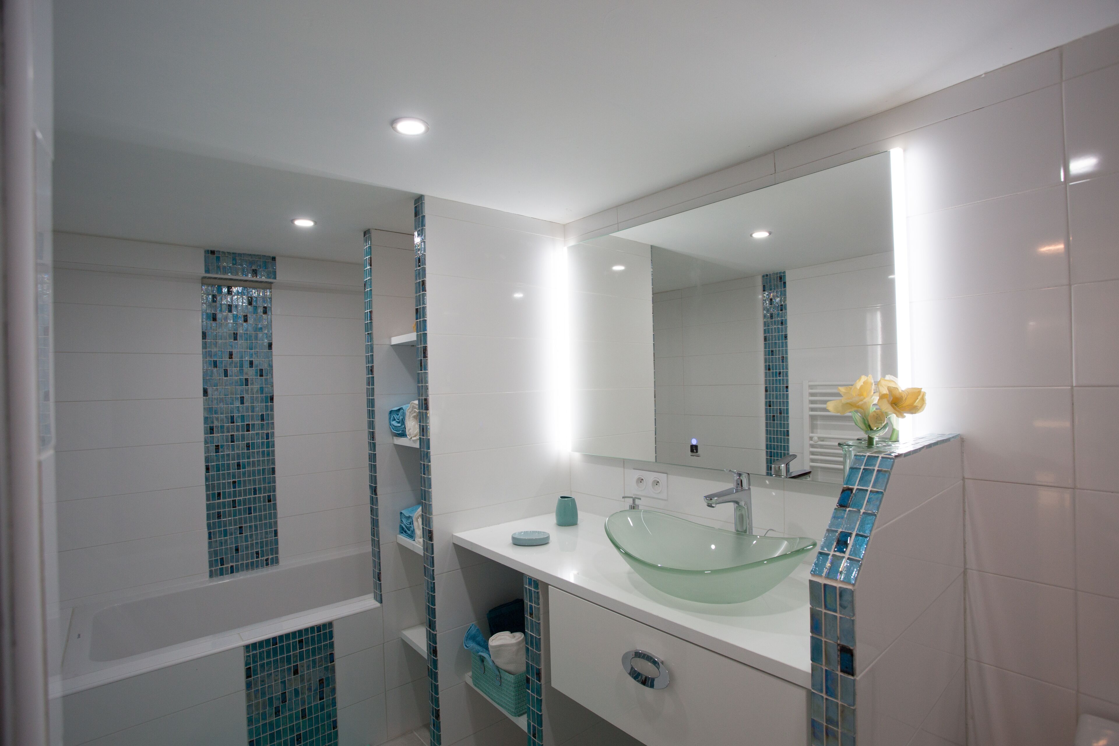 Un baño moderno con un lavabo de vidrio, azulejos blancos y un espejo con un revestimiento de azulejos azules.