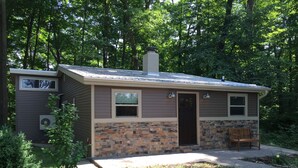 A small cabin with a stone foundation and a sloped roof, surrounded by dense forest.