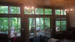 A cozy cabin interior with a large window overlooking a forest, a wooden ceiling, and a black leather couch with patterned cushions.