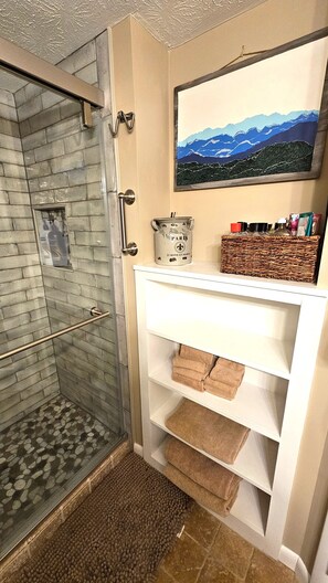 A bathroom with a glass shower, a white shelf with towels, and a framed landscape painting above it.
