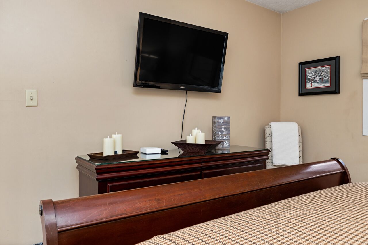 A hotel room with a bed, a dresser, a flat-screen TV mounted on the wall, a framed picture, and a small table with candles.