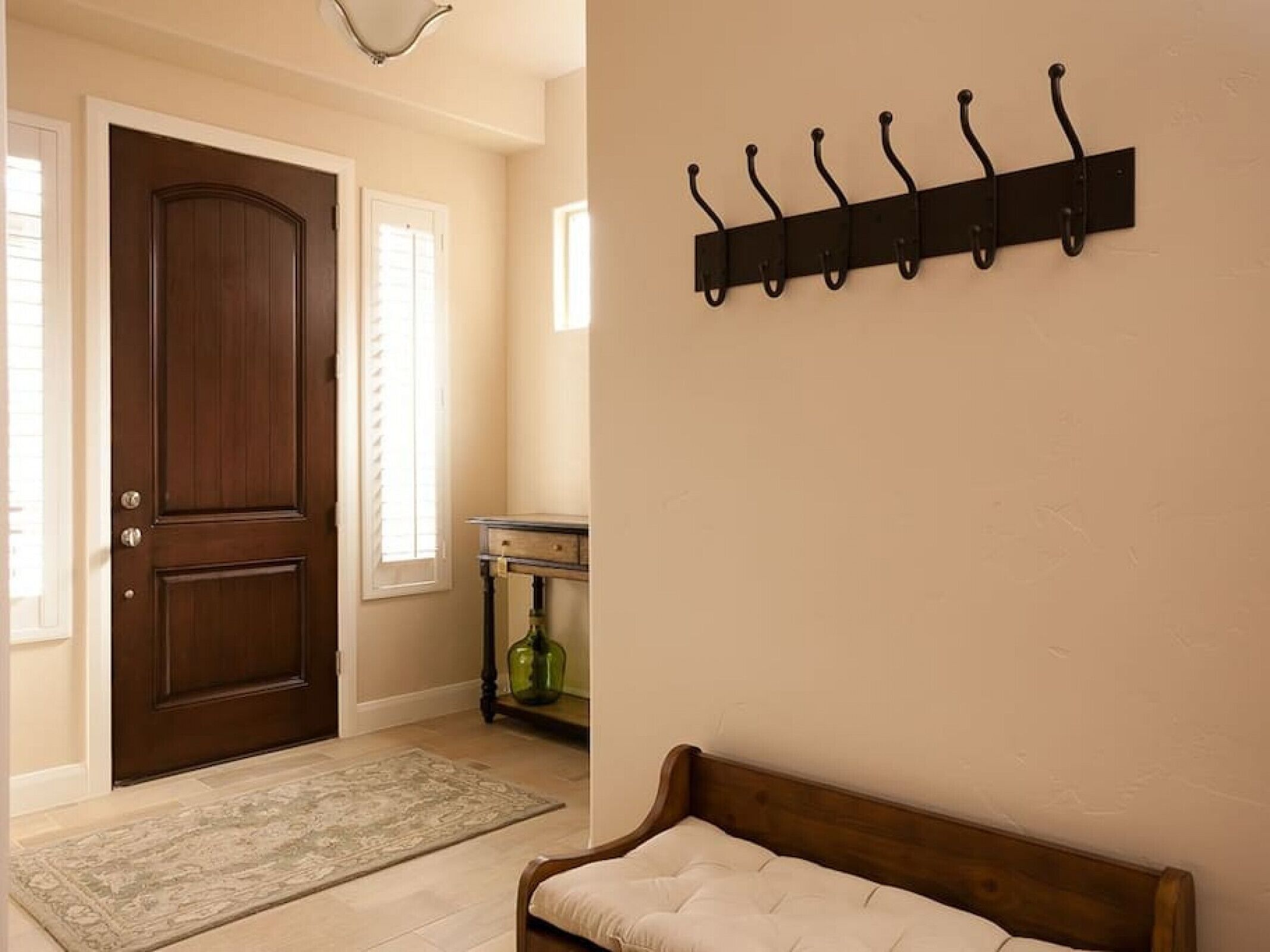 Un pasillo con una puerta de madera, un banco, un perchero en la pared y una mesita con un jarrón verde.