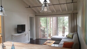 A modern living room with a flat-screen TV on a white shelf, a wooden coffee table with a magazine, a grey sofa with orange cushions, and a large sliding glass door leading to a forested area.