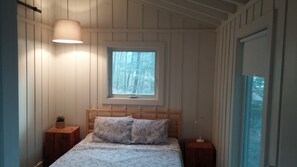 A bedroom with a wooden bed, two bedside tables, a window with a view of trees, and a hanging light fixture.