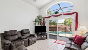 A spacious living room with a large flat-screen TV, two reclining sofas, and a sliding glass door leading to a balcony with a pool. - 4BR Disney Oasis: Private Screened Pool + FREE Pool Heat! (20 Min to Parks) (Kissimmee)