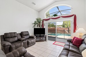 A spacious living room with a large flat-screen TV, two reclining sofas, and a sliding glass door leading to a balcony with a pool.
