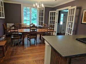 A dining area with a large wooden table and chairs, a kitchen island with a granite countertop, and a view of the outdoors through large windows. - Stonehouse w/ Hot Tub, Walk to GAP Trail & Town (Confluence)