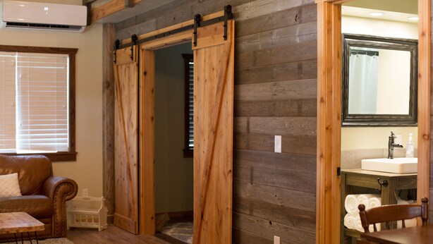 Une pièce avec un plancher en bois, un canapé en cuir marron, une table en bois, une porte coulissante de style grange, une salle de bain avec un miroir et un lavabo, et une fenêtre avec des stores.