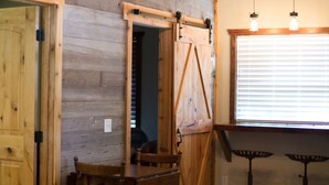 Un intérieur rustique avec des murs en bois, une table à manger entourée de chaises, un lustre et une grande fenêtre équipée de stores.