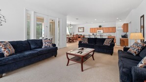 Ein geräumiges Wohnzimmer mit zwei dunklen Sofas, einem Couchtisch und gemusterten Kissen. Im Hintergrund ist eine Küche mit Holzschränken und ein Essbereich mit Tisch und Stühlen zu sehen.