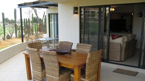 A dining area with a wooden table and wicker chairs, a glass door leading to a living area, and a view of cacti and a garden.