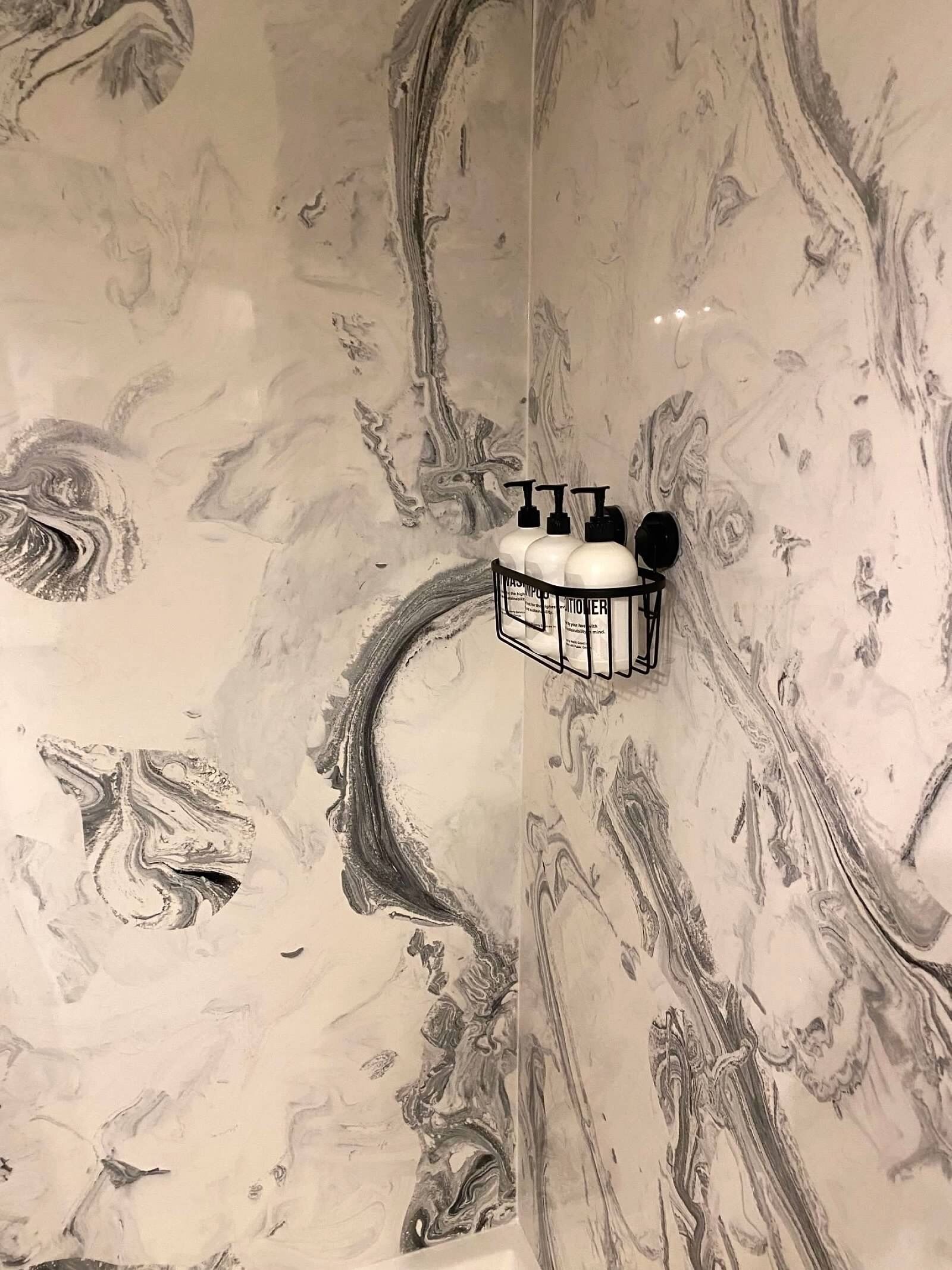 Une salle de bain avec un mur en marbre orné d’un motif tourbillonnant. On y trouve un distributeur de savon noir et un récipient noir, tous deux posés sur un support blanc.