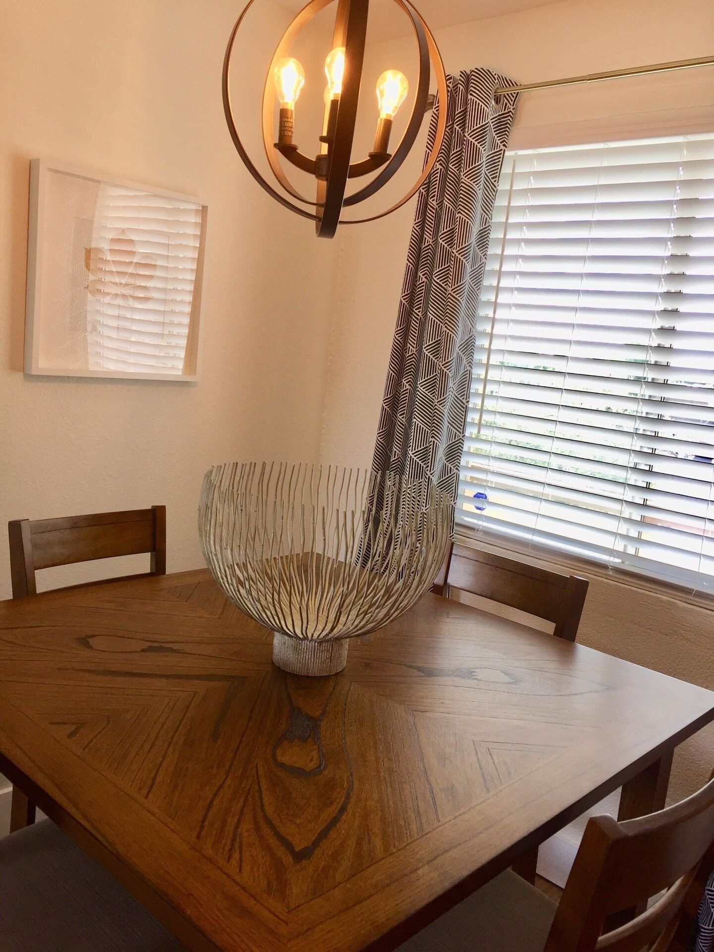 Una mesa de comedor con un centro de mesa tejido, sillas de madera, una lámpara de pared y una ventana con persianas.
