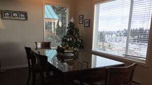 A dining area with a glass-top table, wooden chairs, a Christmas tree, and a view of a snowy landscape through large windows. - Ski In/out For Both Downhill And Cross-country (Vernon)