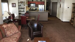 Una sala de estar con un sofá, una mesa de centro y un comedor con sillas y una mesa. Se observa una zona de cocina con armarios y un refrigerador.