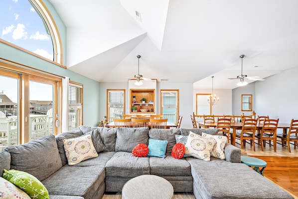 A spacious living room with a sectional sofa, dining table, and chairs, large windows, and a skylight.