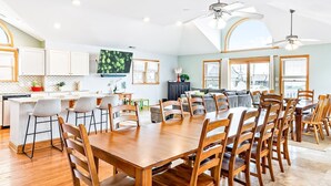 A spacious dining area with a long wooden table and chairs, a kitchen area with white cabinets, and a ceiling fan. - Semi-Oceanfront, Heated Pool and Hot Tub! (Nags Head)