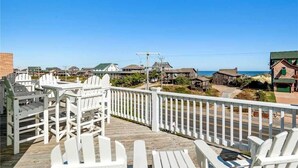 A wooden deck with white Adirondack chairs overlooking a beach with several buildings. - Semi-Oceanfront, Heated Pool and Hot Tub! (Nags Head)
