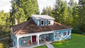 A two-story house with a sloped roof, a front porch with chairs, and a red door.