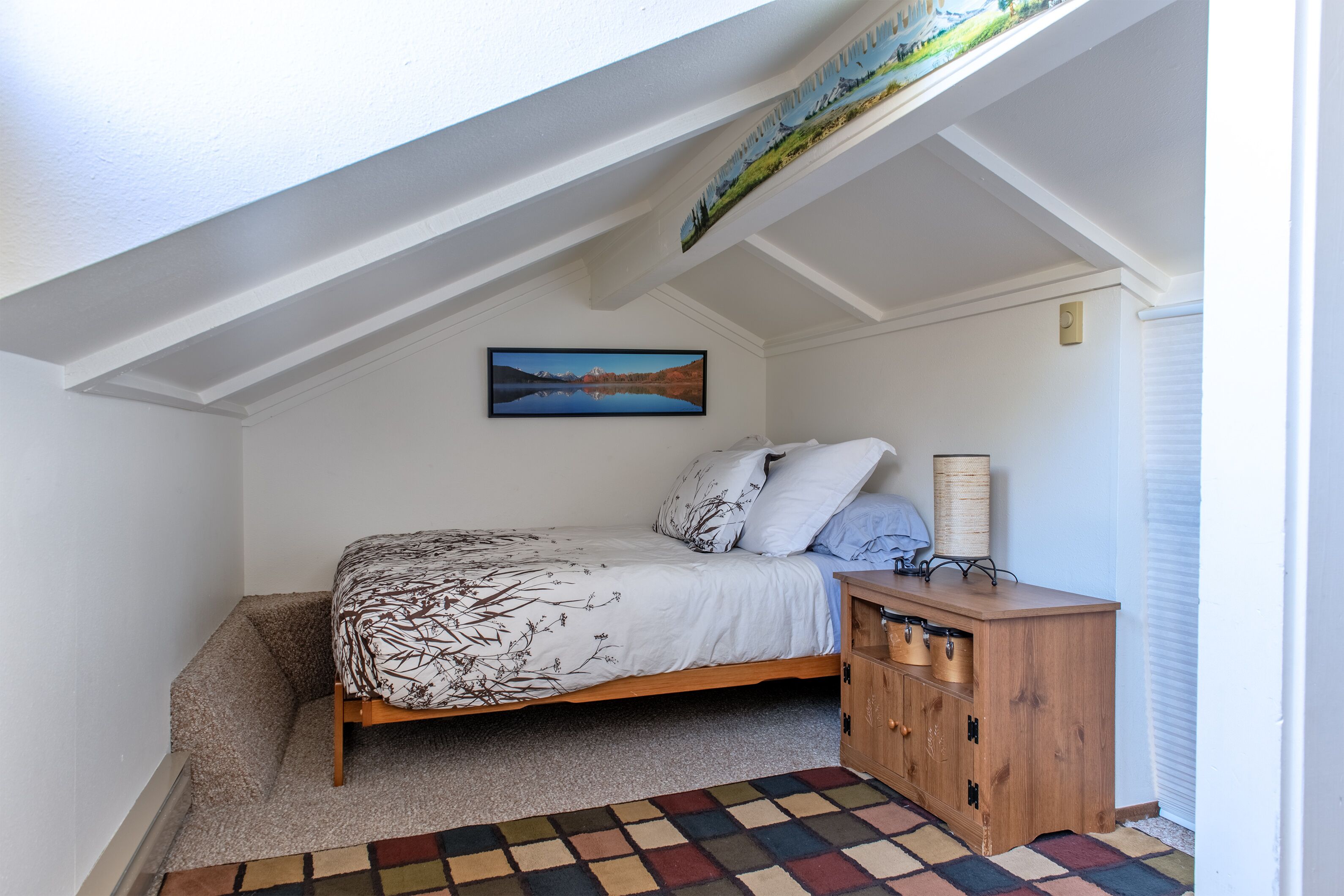 Een slaapkamer met een schuine plafond, een bed met beddengoed met een patroon, een nachtkastje met een lamp en een schilderij aan de muur.