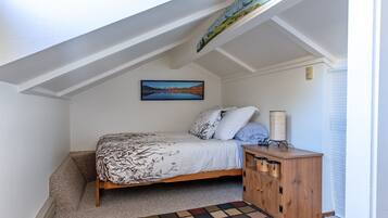 Een slaapkamer met een schuine plafond, een bed met beddengoed met een patroon, een nachtkastje met een lamp en een schilderij aan de muur.