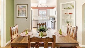 Uma área de refeições com mesa e cadeiras de madeira, uma planta central e uma cozinha ao fundo.