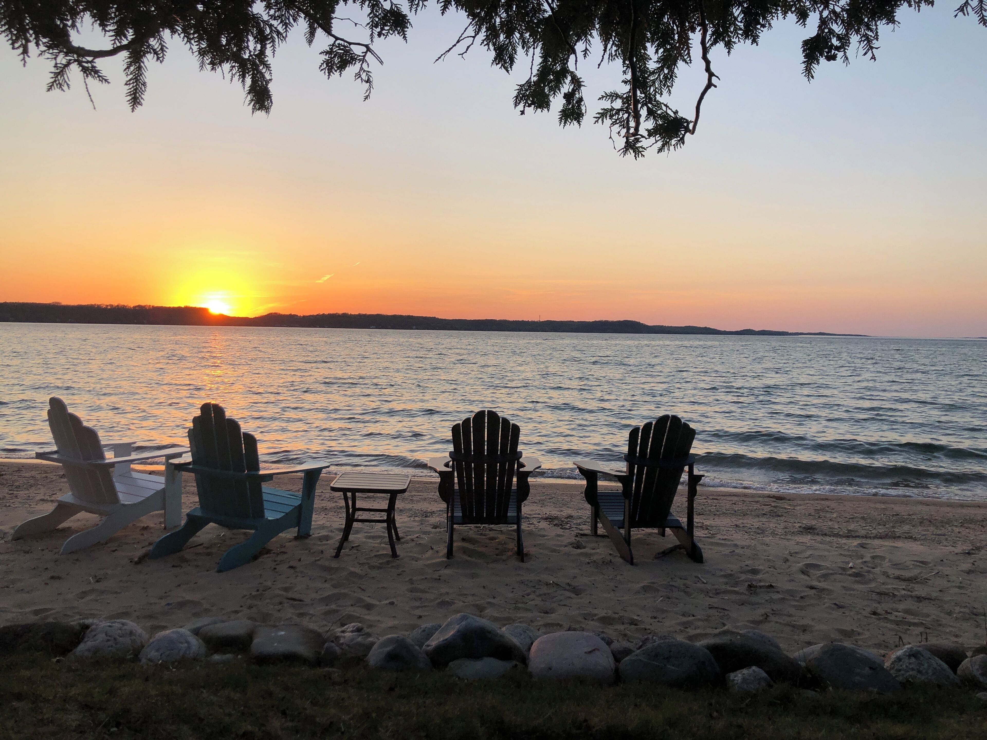 A beach with three Adirondack chairs and a small table facing the sunset over water.
