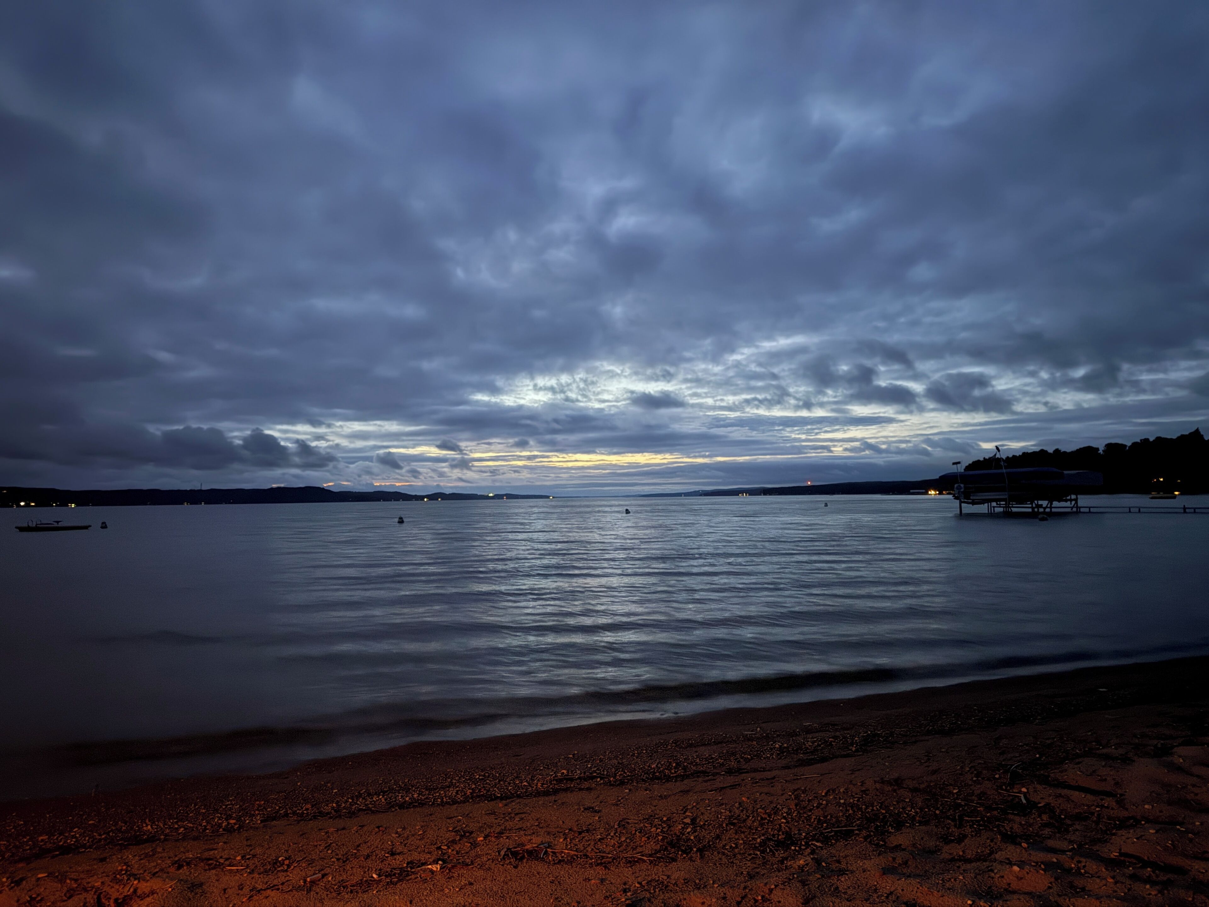 A beach with calm waters, a distant shoreline, and a cloudy sky.