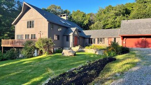 Une maison à deux étages dotée d’une terrasse en bois, de fondations en pierre et d’une allée gravillonnée.