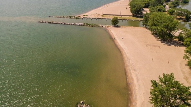Ovenudblik over en strand med sandet bred, en bro, der strækker sig ud i vandet, og en række træer langs kystlinjen.