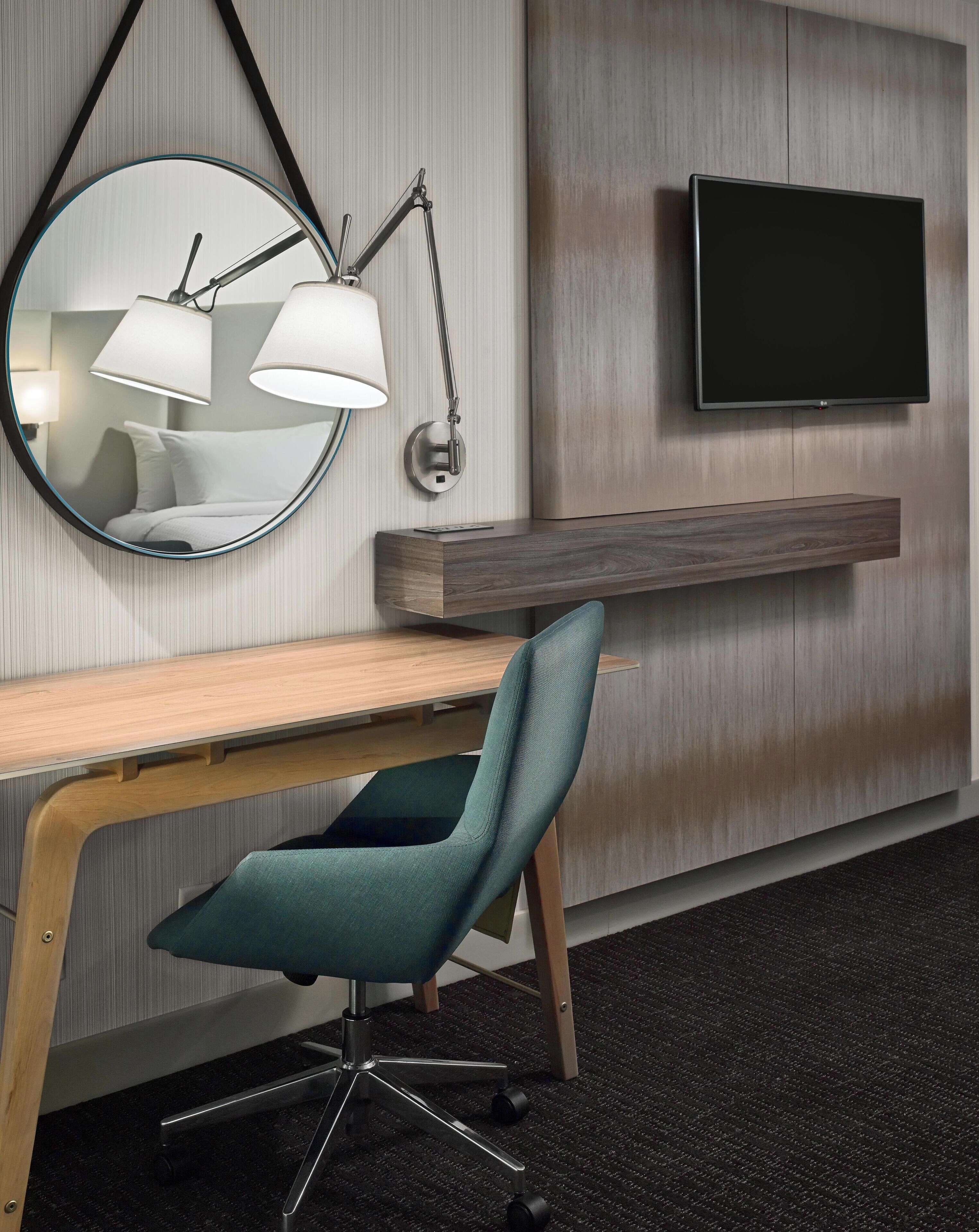 Ett modernt hotellrum med ett skrivbord, en stol, en väggmonterad spegel, en väggmonterad TV och en liten hylla.