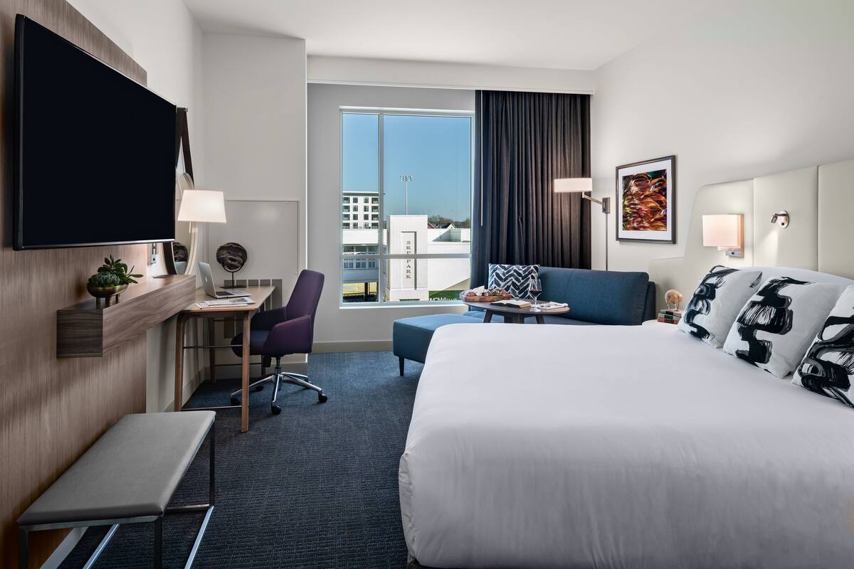 Ein modernes Hotelzimmer mit einem großen Bett, einem Schreibtisch mit Computer, einem Sessel, einem kleinen Tisch und einem großen Fenster mit Blick auf Gebäude.