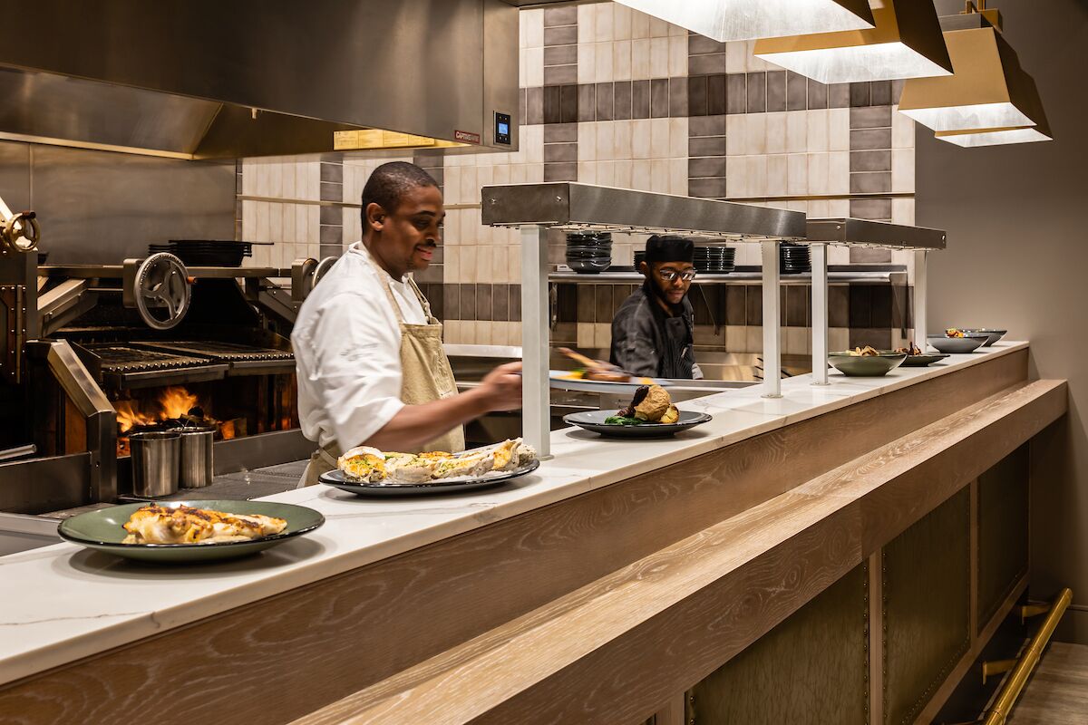 A professional kitchen with chefs preparing food, a wood-paneled counter, and stainless steel appliances.