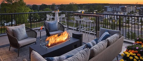 A balcony with a fire pit, outdoor seating, and a view of the cityscape at sunset.