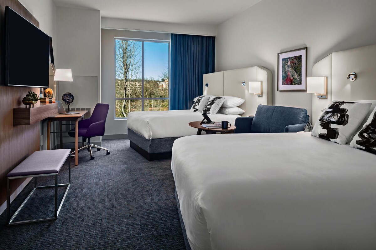 A hotel room with two beds, a desk, a chair, a TV, and a window with a view.