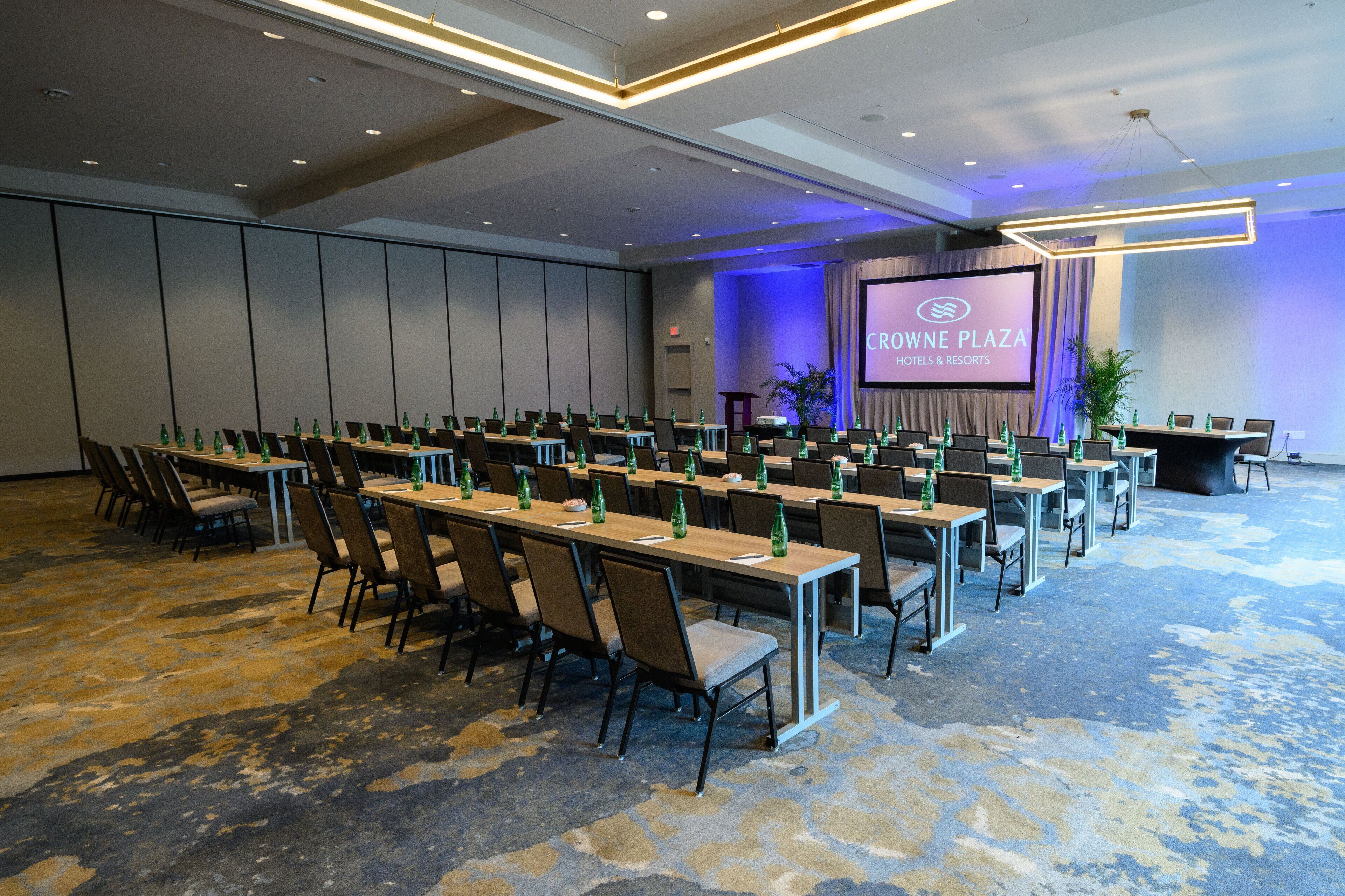 A conference room with rows of tables and chairs, a large screen displaying 'CROWNE PLAZA HOTEL & RESORTS', and a carpeted floor.